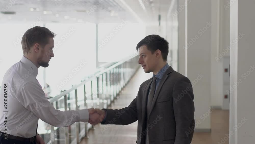 Two young businessmen shaking hands in office gallery, slow motion. Concept: people, business, bargain Two young businessmen shaking hands in office gallery, slow motion. Concept: people, business, bargain