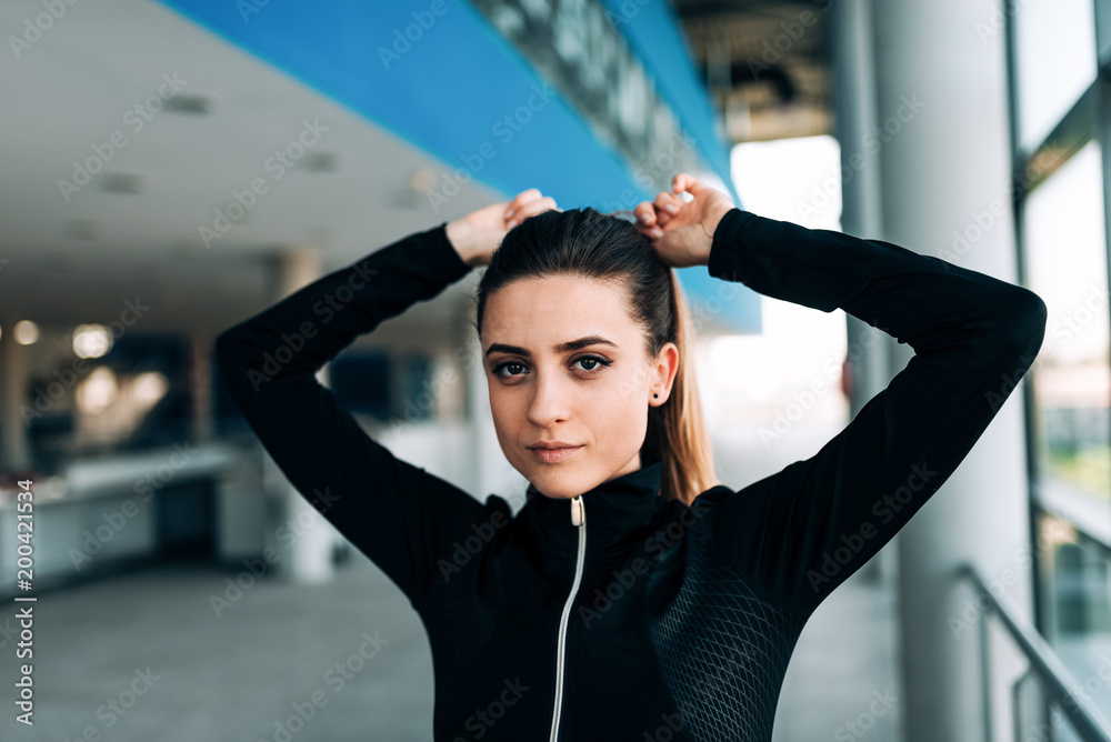 Close up portrait of beautiful woman in black tracksuit tying her hair before workout.