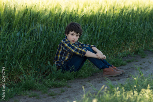 A sad boy in jeans and a shirt is sitting in the field.
