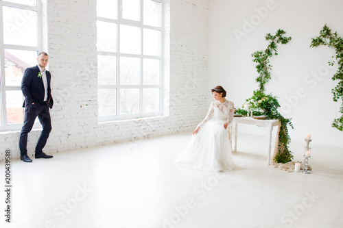 The beautiful bride is seated on the chair in the studio. The bridesmaid in the suit stands at the window