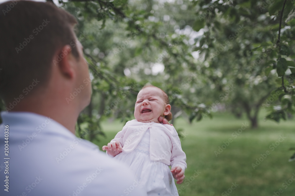The father holds a crying newborn girl in a summer green Park. A man stands with his back to the camera. The father holds a crying newborn girl in a summer green Park. A man stands with his back to the camera.