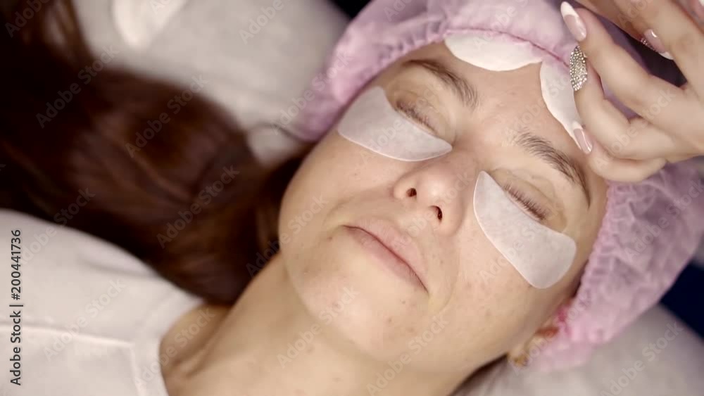 well groomed woman is in a procedure of eyelashes lamination, lying face up and master is applying keratin composition well groomed woman is in a procedure of eyelashes lamination, lying face up and master is applying keratin composition