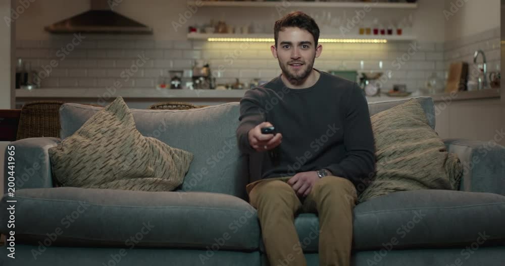 Young, attractive man sits down on a couch and turns on the television using a remote control.