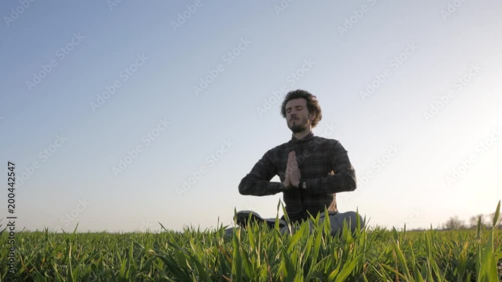 Young man in casual wear relaxing in green field after work, meditation of male against sunset or sunrise Young man in casual wear relaxing in green field after work, meditation of male against sunset or sunrise