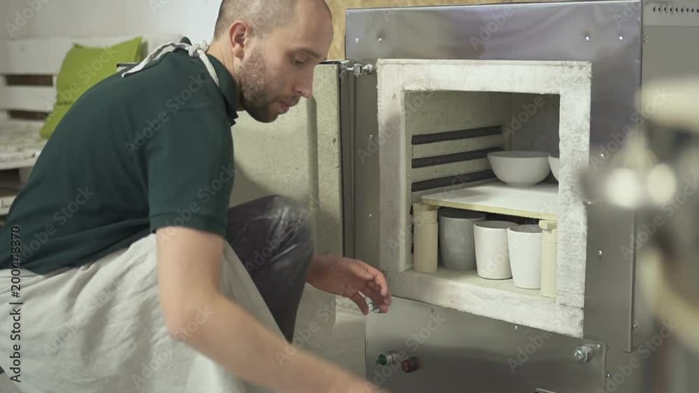 In the pottery workshop man puts clay pots for firing in the oven. Bearded craftsman in an apron places a billet in hot stove to create ceramic dishes. In the pottery workshop man puts clay pots for firing in the oven. Bearded craftsman in an apron places a billet in hot stove to create ceramic dishes.
