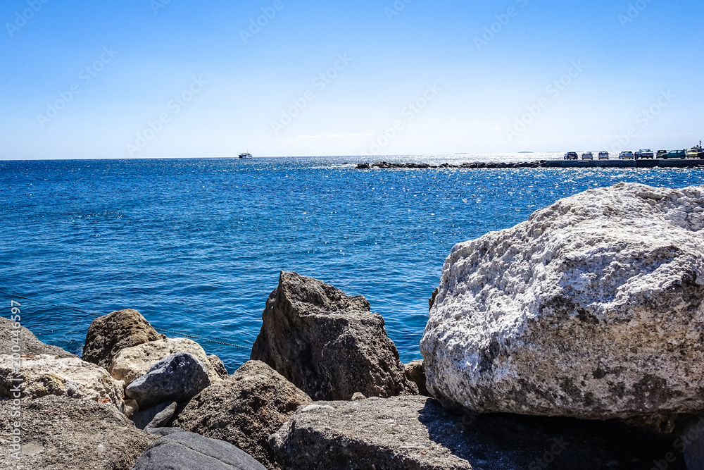 beautiful Mediterranean view on a summer sunny day, blue clear sky and blue water, with shore stones in the forefront, a boat in the distance.