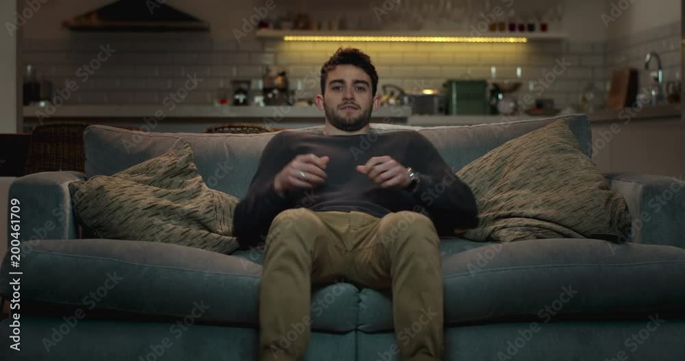 Man walks into the living room and sits on a couch to watch tv