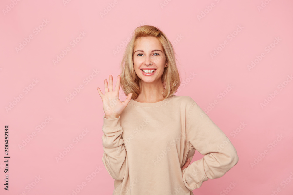 Hey! Young pretty woman greet her friends and feels happy to meet them. Blonde girl smiles and shows her palm. Isolated over pink background Hey! Young pretty woman greet her friends and feels happy to meet them. Blonde girl smiles and shows her palm. Isolated over pink background