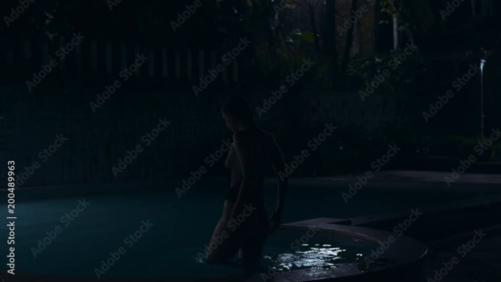 Night footage of beautiful young girl with amazing athletic sexy body in black bikini comes into the water and swims in the swimming pool with light, old vintage wall. Movie shooting of female model