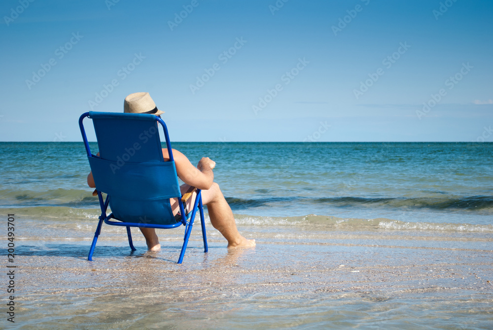 young man sitting on a deckchair on the sea shore ocean resting sunbathing looking at the sea beach shore sand sky summer rest weekend