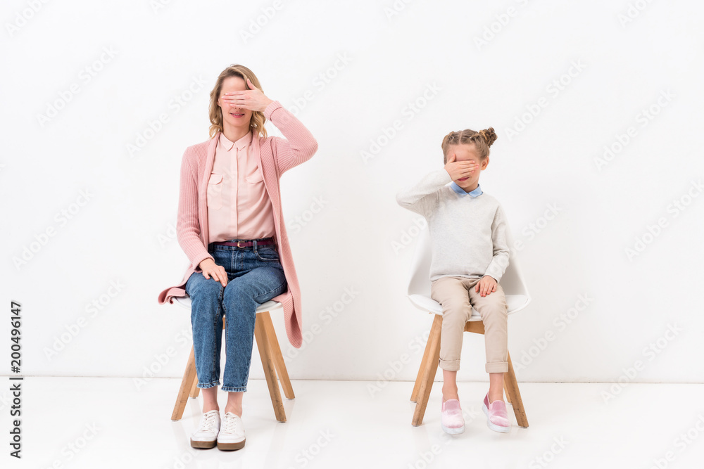 mother and daughter sitting on chairs and covering eyes with hands on white