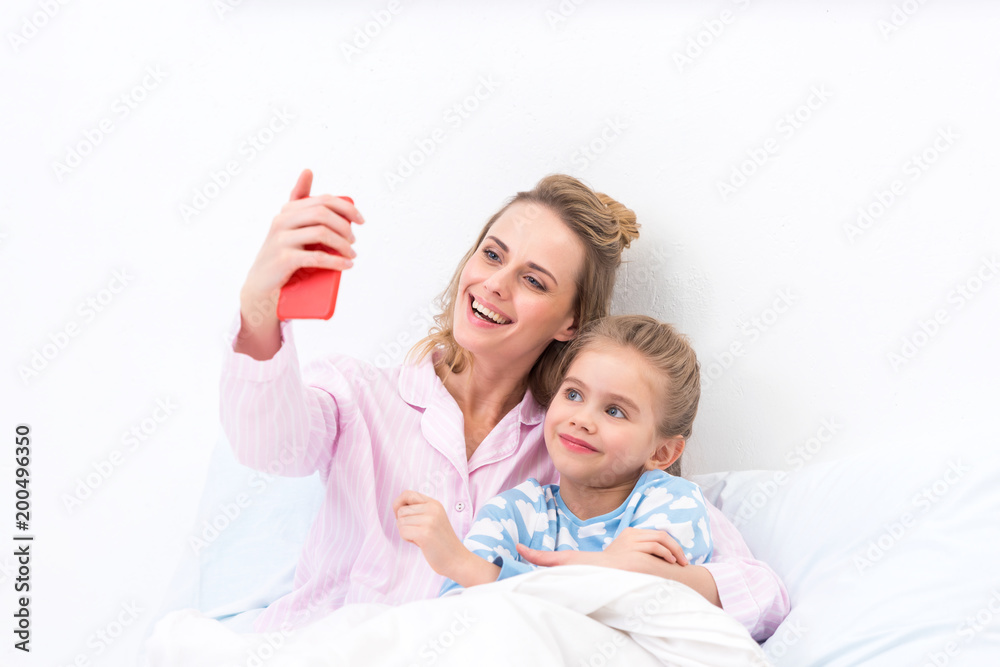 smiling mother and daughter taking selfie with smartphone on bed at home smiling mother and daughter taking selfie with smartphone on bed at home