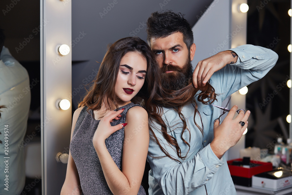 Happy positive woman looks at her hair makeup artist and bearded man  hairdresser in the salon. Serious brutal man hairdresser making hairstyle  for pretty woman in barber shop. Beautiful fashion couple Stock