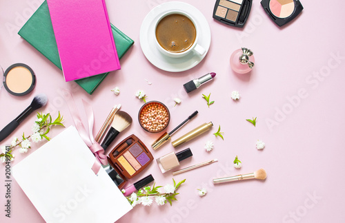 Various cosmetic products for makeup and white spring flowers in a white gift bag, notepad, cup of coffee on a pink pastel background top view with copy space photo