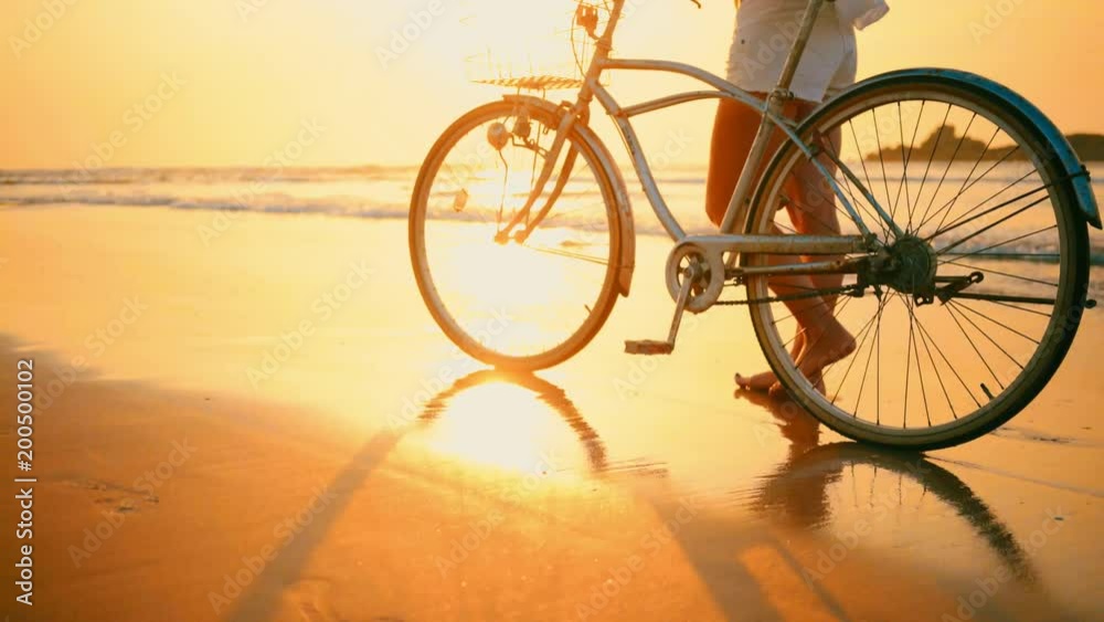 Woman in her hat walking with bicycle along beach sand at summer time under burning sunset sky, lifestyle carefree Woman in her hat walking with bicycle along beach sand at summer time under burning sunset sky, lifestyle carefree
