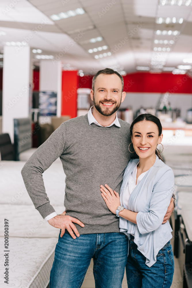 smiling couple looking at camera in furniture store with arranged mattresses smiling couple looking at camera in furniture store with arranged mattresses