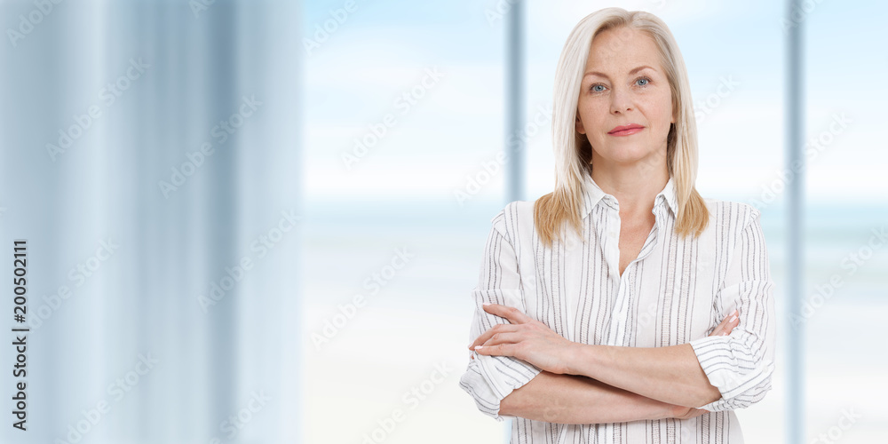 Attractive middle aged business woman with folded arms is standing near the window. Summer landscape outside the window as a background. Attractive middle aged business woman with folded arms is standing near the window. Summer landscape outside the window as a background.