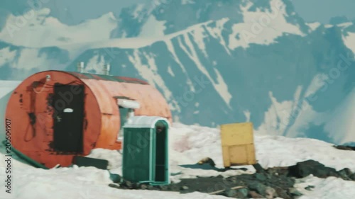 Orange metal drum tourist shelter with green biotoilet at hight rocky mountain covered in snow scenic landscape on sunny day with blue sky, tracking shot photo