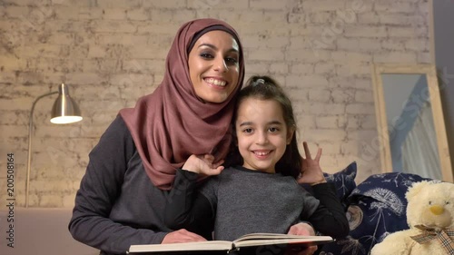 Young woman in hijab smiling and sits on sofa with her daughter and teaches her how to read, book, happy family concept, smile and look at the camera, wave their handshome comfort in the background 50 photo