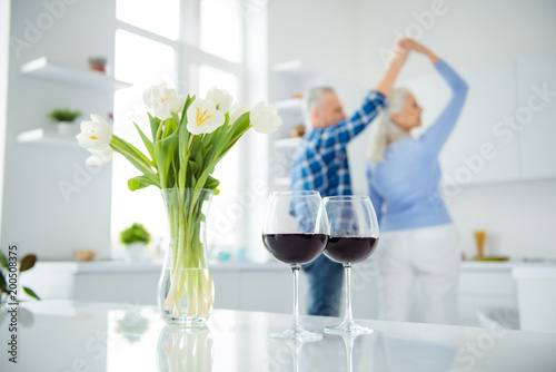 Fototapeta Naklejka Na Ścianę i Meble -  Portrait of lovely happy modern stylish attractive couple, dancing in the kitchen on blurred background, two glasses of red wine, bouquet of tulips in vase standing on the table, spring time