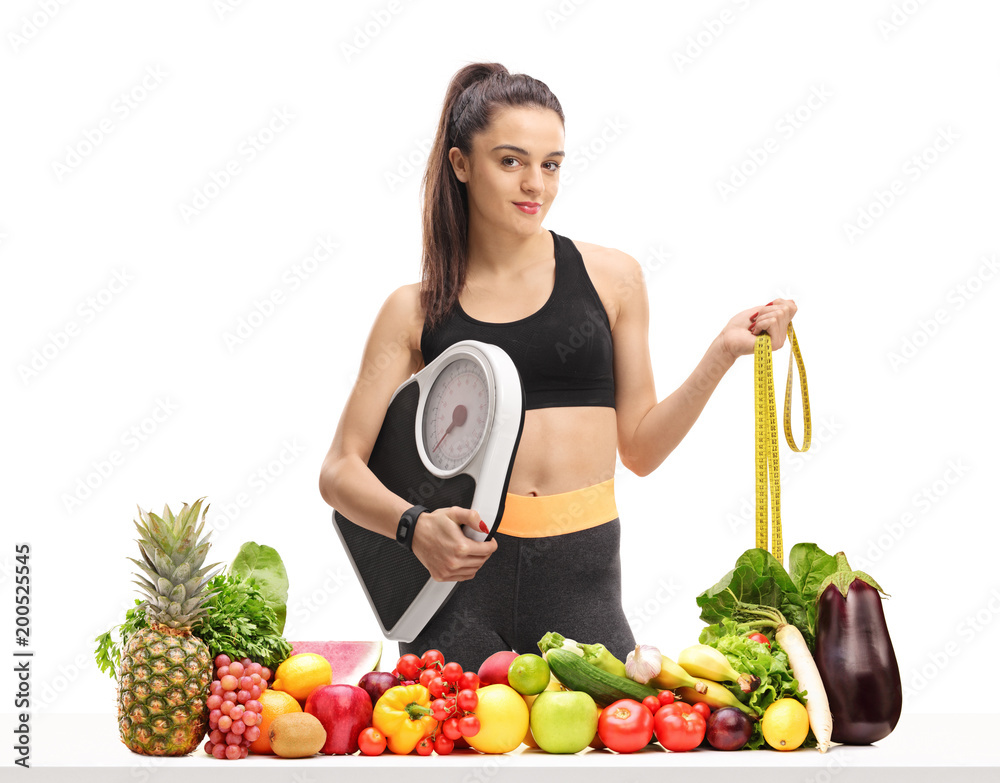 Fitness woman holding a weight scale and a measuring tape behind a table with fruit and vegetables Fitness woman holding a weight scale and a measuring tape behind a table with fruit and vegetables