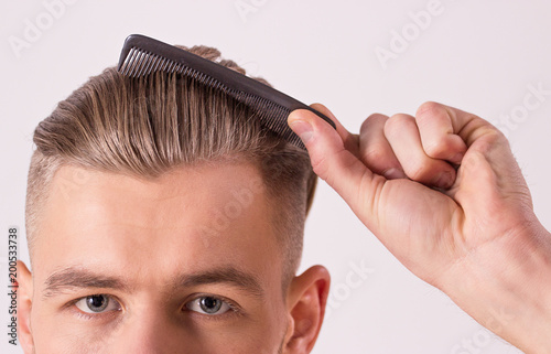 Close up of young attractive man brushing hair with comb in hand, isolated on white background. Man's face with modern and stylish haircut. Male beauty, hair style. Hipster combing hair or making hair photo