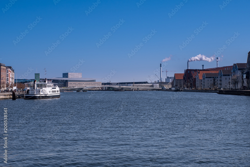 A view of the Inderhavnsbroen in Copenhaven, Denmark, and a moored up ship on the left. Towers belching out white smoke are on the right.