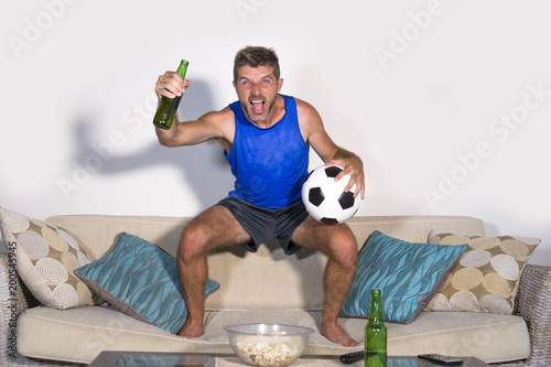 young attractive man happy and excited watching football match on TV celebrating victory goal jumping on sofa couch with beer and popcorn © TheVisualsYouNeed