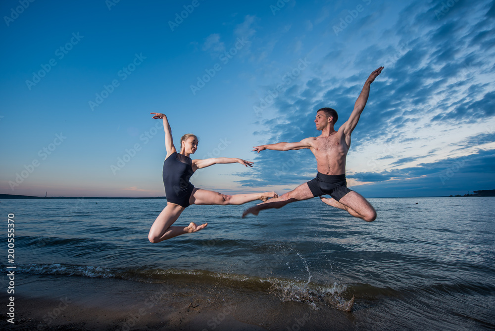 Young guy and girl in black swimwear high jump and dance on the beach in the evening. Street ballet.