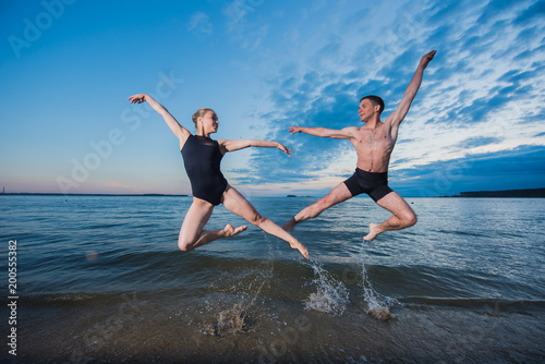 Young guy and girl in black swimwear high jump and dance on the beach in the evening. Street ballet.
