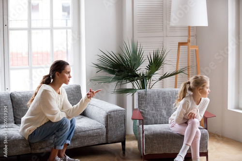 Bild auf Leinwand Young strict mother or sister scolding stubborn sulky kid girl in living room at