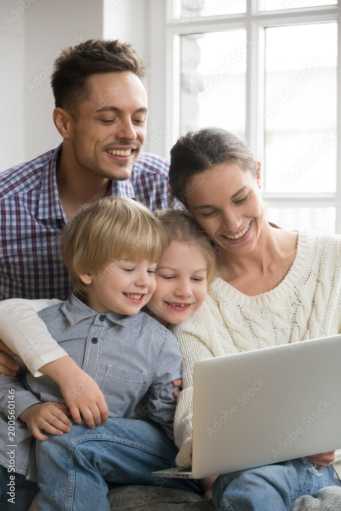 Happy family with adopted kids having fun using laptop together sitting on sofa, parents and son daughter relaxing at home with computer, smiling couple with children watching video online, vertical Happy family with adopted kids having fun using laptop together sitting on sofa, parents and son daughter relaxing at home with computer, smiling couple with children watching video online, vertical