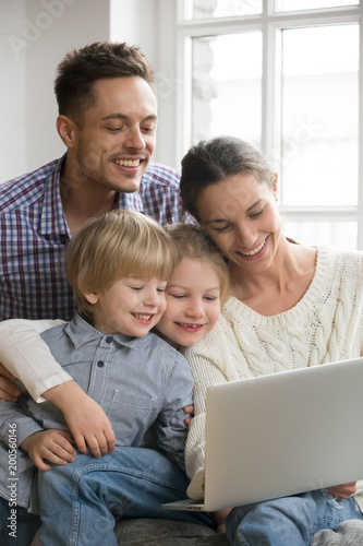 Happy family with adopted kids having fun using laptop together sitting on sofa, parents and son daughter relaxing at home with computer, smiling couple with children watching video online, vertical