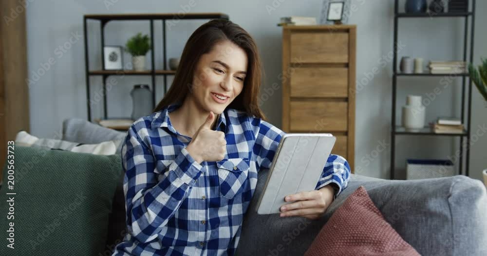 Young brunette pretty woman smiling in the camera, showing gestures and talking while videochatting on the tablet in the living room. Indoor Young brunette pretty woman smiling in the camera, showing gestures and talking while videochatting on the tablet in the living room. Indoor