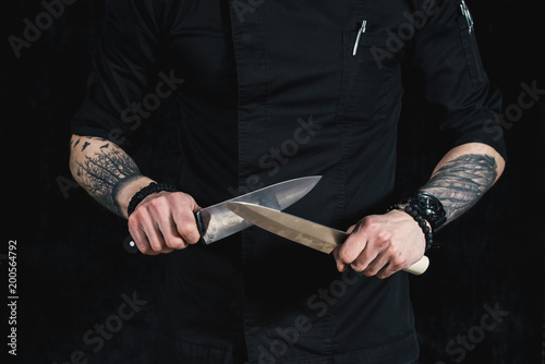 Young attractive man chef in black uniform hands in tattoos holds two knives. black background