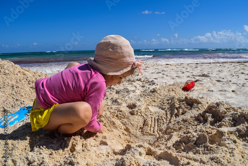 Kleines Mädchen mit rosa farbenem Sonnenhut spielt mit Sand am Strand, Hintergrund Strand, Meer, Himmel