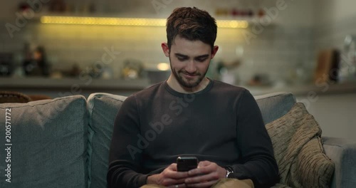 Smiley young man web surfing on his mobile phone on the sofa at home.