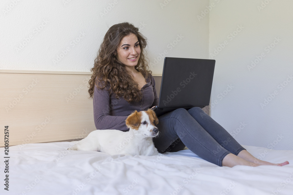 beautiful young woman sitting on bed with her cute small dog besides. She is working on laptop and smiling. Breakfast at home, indoors and lifestyle beautiful young woman sitting on bed with her cute small dog besides. She is working on laptop and smiling. Breakfast at home, indoors and lifestyle