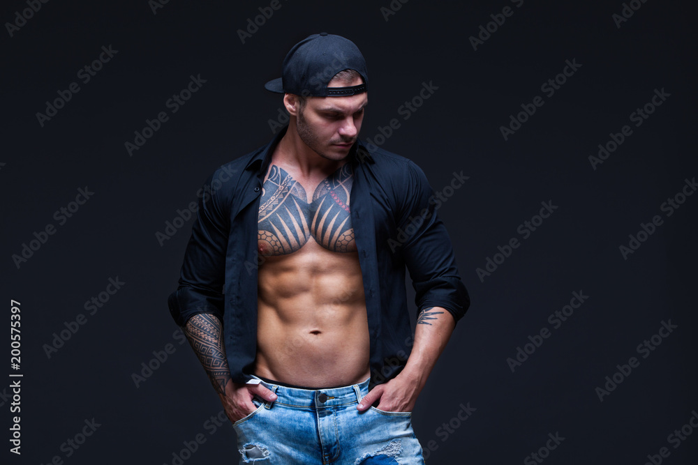 The man dressed blue jeans, black shirt and black baseball cap with tattoos on the dark background. ABS