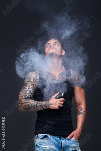 Vaper. The man with a muscular torso with tattoos smoke an electronic cigarette on the dark background © satyrenko
