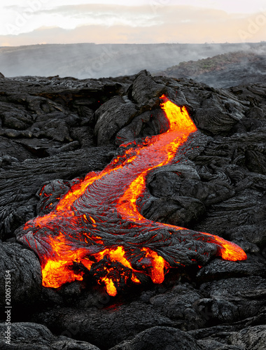 A lava flow emerges from an earth column and flows in a black volcanic landscape, in the sky shows the first daylight - Location: Hawaii, Big Island, volcano 