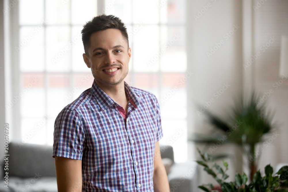 Happy young adult man posing indoors at home in living room, cheerful male student or new real estate owner smiling looking at camera, handsome casual single millennial guy headshot portrait Happy young adult man posing indoors at home in living room, cheerful male student or new real estate owner smiling looking at camera, handsome casual single millennial guy headshot portrait