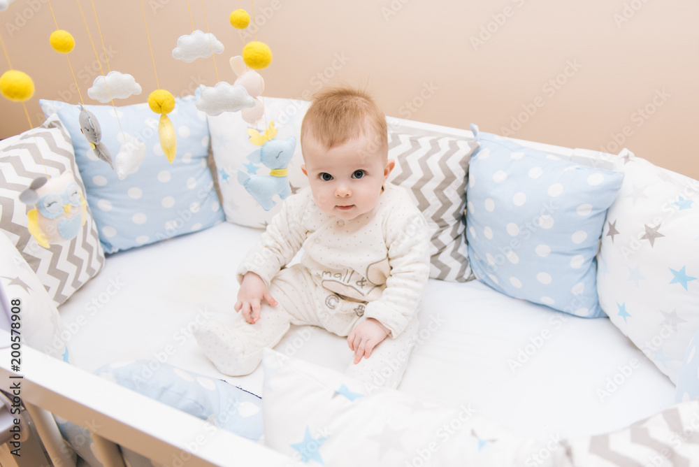 Cute baby sitting in a white round bed. Light nursery for young children. Toys for infant cot. Smiling child playing with mobile of felt in sunny bedroom. Cute baby sitting in a white round bed. Light nursery for young children. Toys for infant cot. Smiling child playing with mobile of felt in sunny bedroom.
