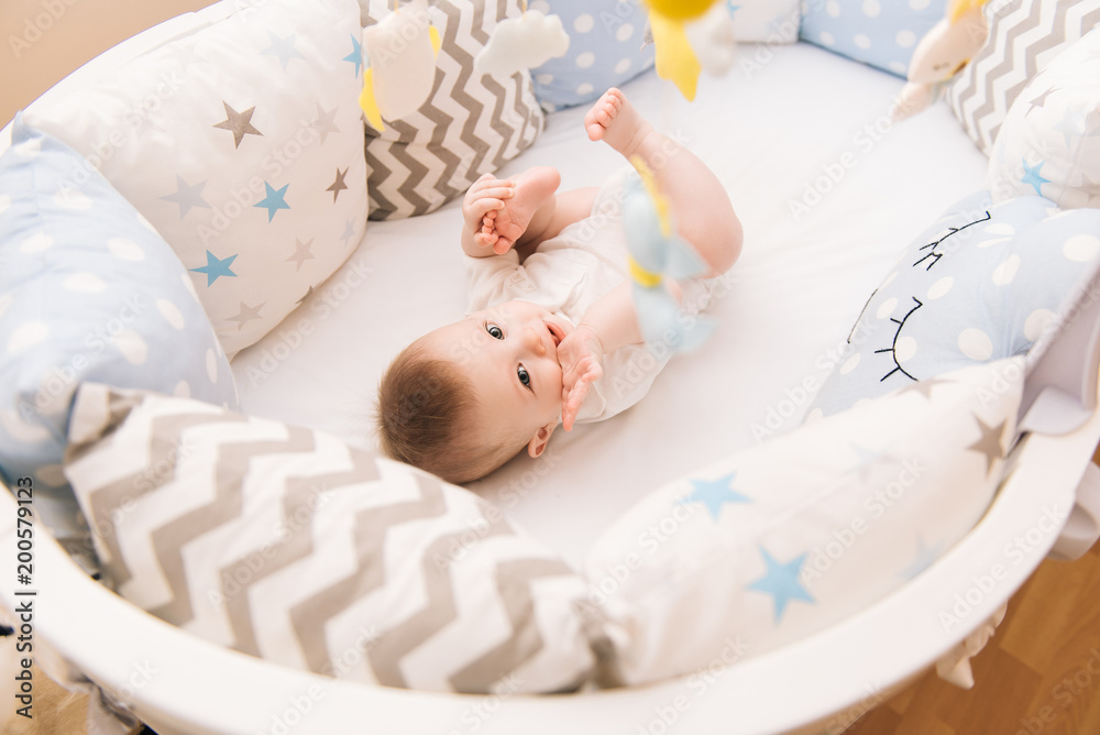 Cute baby lies in a white round bed. Light nursery for young children. Toys for infant cot. Smiling child playing with mobile of felt in sunny bedroom.