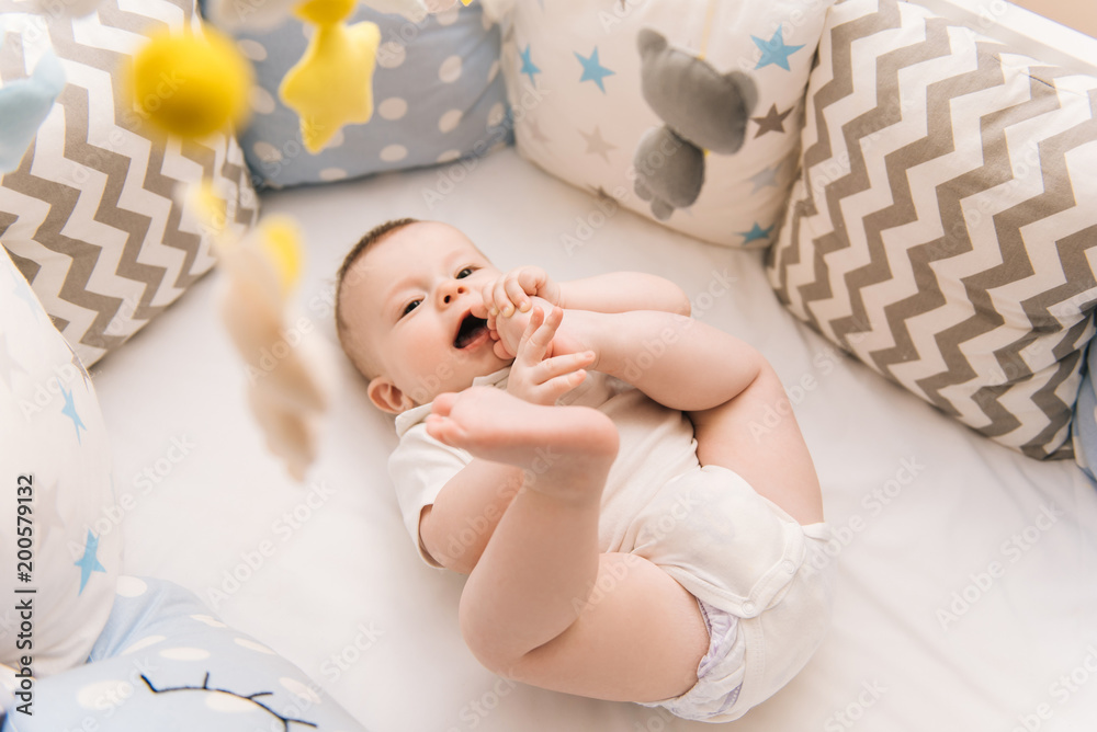 Cute baby lies in a white round bed. Light nursery for young children. Toys for infant cot. Smiling child playing with mobile of felt and chewing his feet in sunny bedroom.