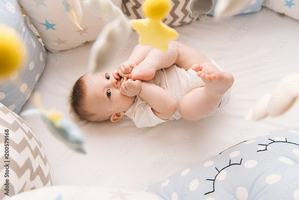 Cute baby lies in a white round bed. Light nursery for young children. Toys for infant cot. Smiling child playing with mobile of felt and chewing his feet in sunny bedroom.