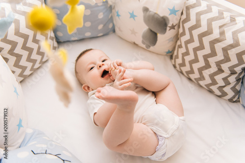 Obraz na płótnie Cute baby lies in a white round bed. Light nursery for young children. Toys for infant cot. Smiling child playing with mobile of felt and chewing his feet in sunny bedroom. w salonie