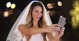 Young bride in wedding dress and veil taking a selfie with cell phone outside