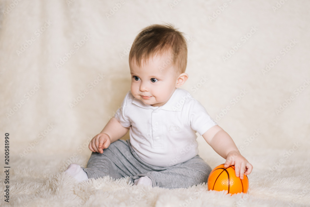 Fototapeta premium Cute young baby sitting on the floor at home and playing with a basketball, sports, healthy and active lifestyle