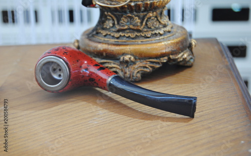 Cachimbo vermelho com traços pretos, pousado numa mesa junto do pé de um cadeeiro em madeira trabalhada  photo